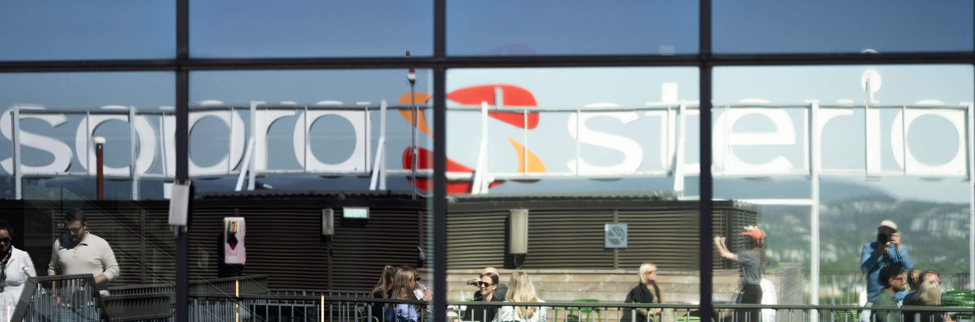 Gjenskinn i vindu av takterrasse med mennesker. I bakgrunnen er et skilt med Sopra Steria-logoen.